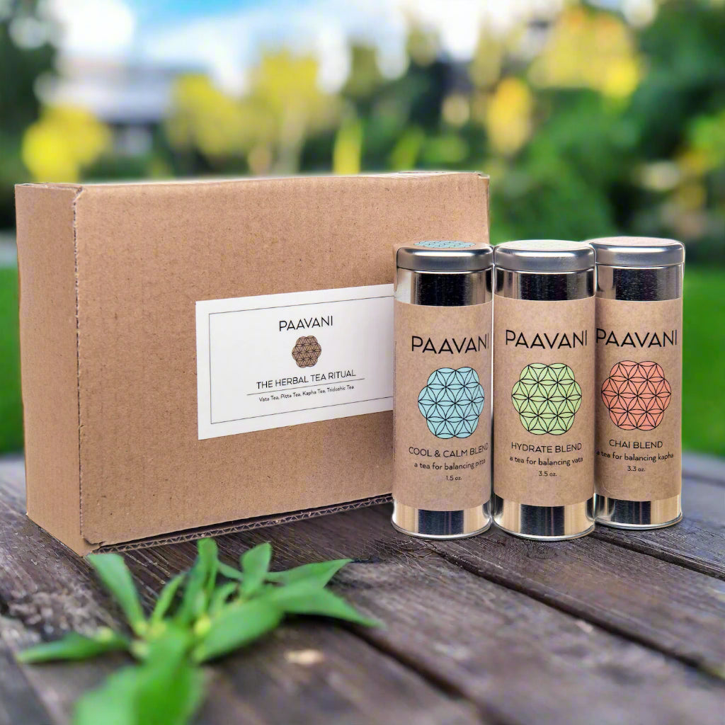 Three metal canisters of Ayurvedic loose-leaf tea blends on a warm linen cloth surrounded by herbs, petals, and roots