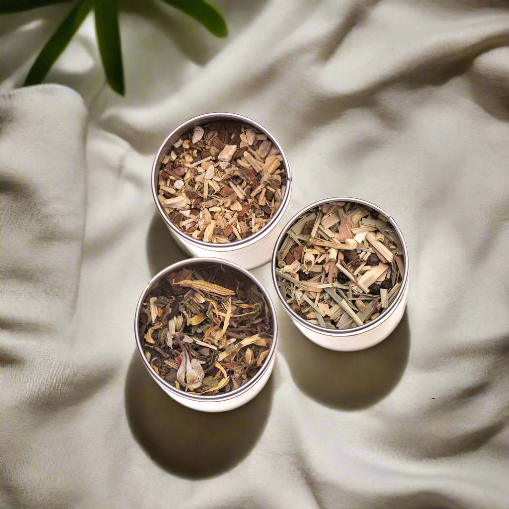 Three metal canisters of Ayurvedic loose-leaf tea blends on a warm linen cloth surrounded by herbs, petals, and roots