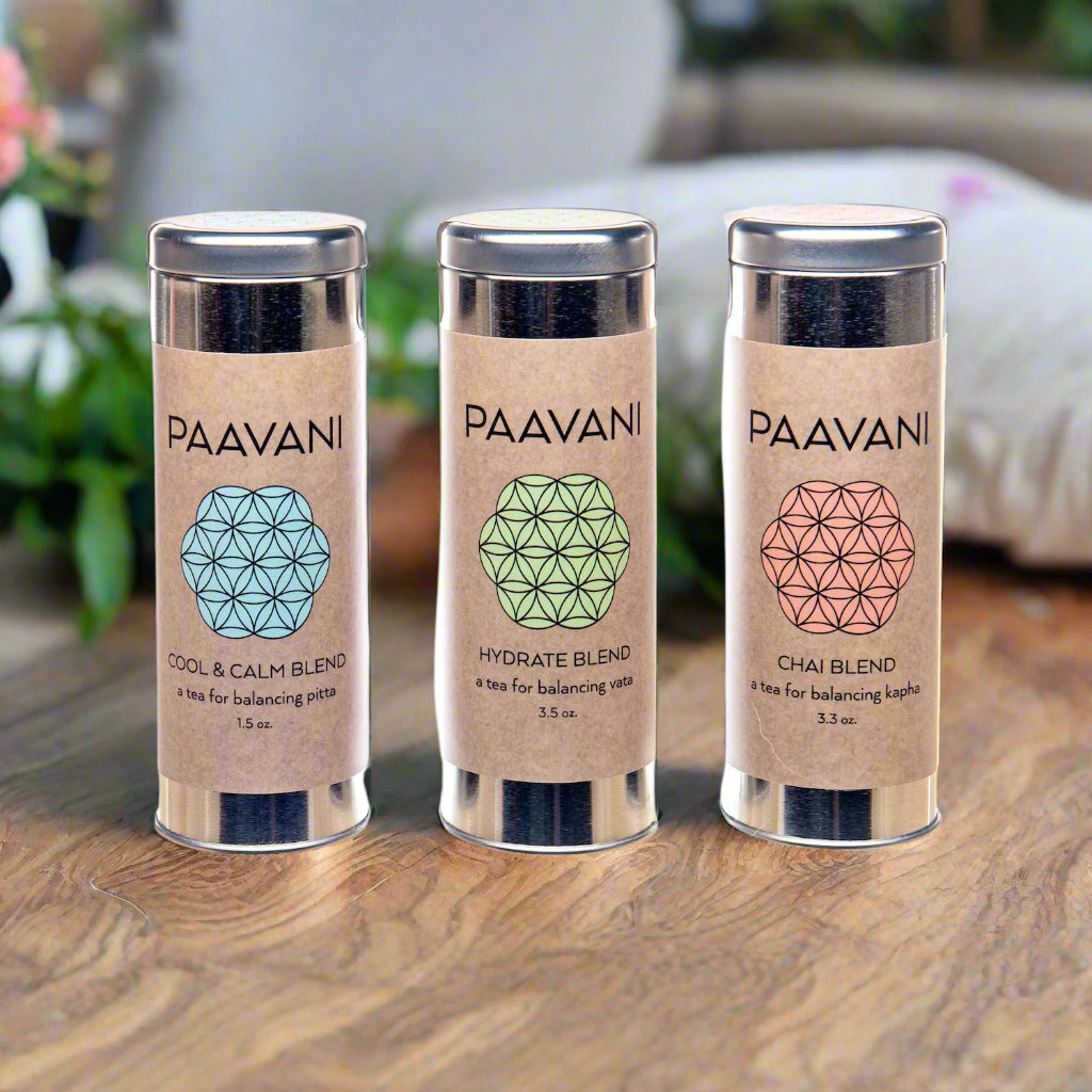 Three metal canisters of Ayurvedic loose-leaf tea blends on a warm linen cloth surrounded by herbs, petals, and roots