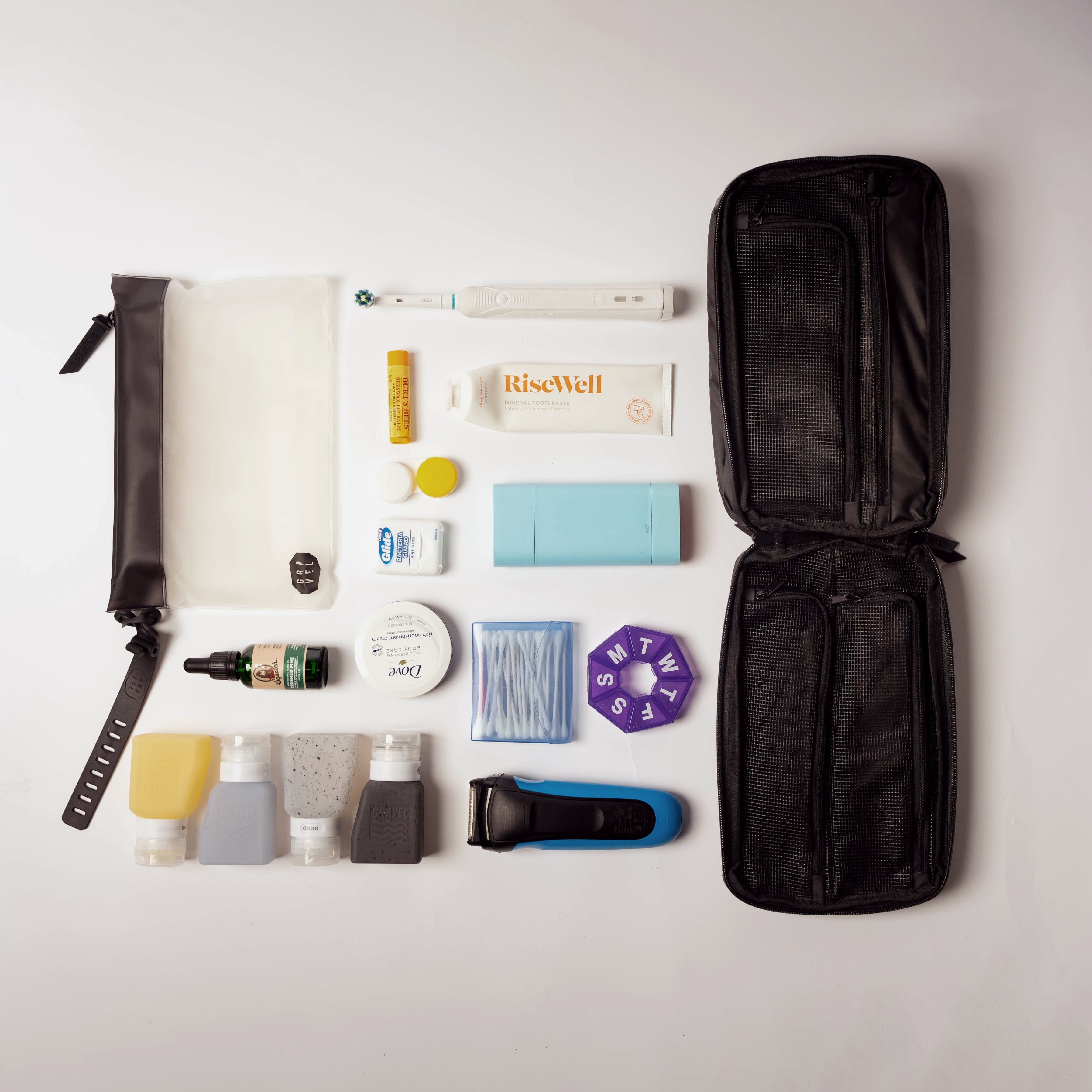 Travel-sized toiletries and grooming items laid out on a white surface with an open black travel bag.