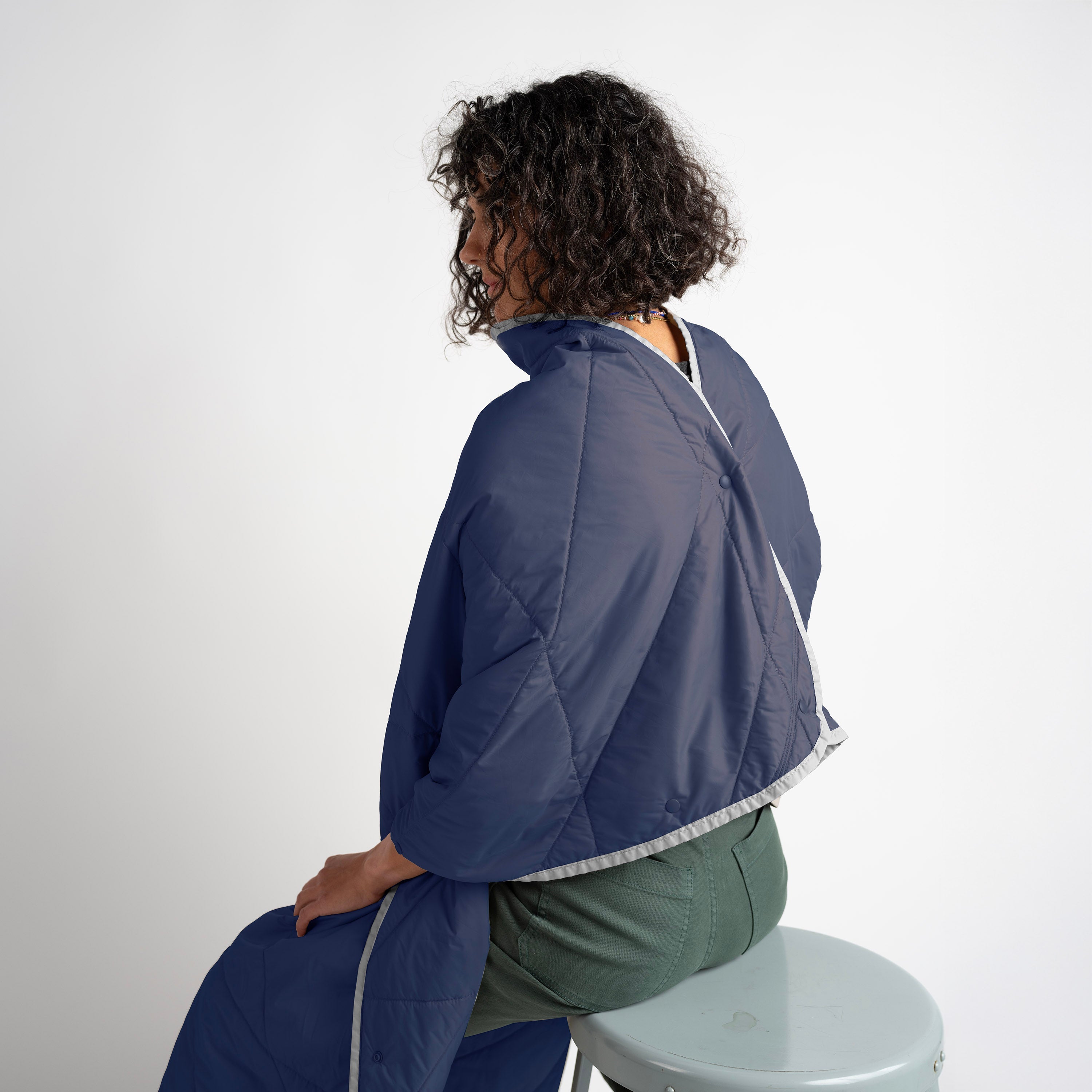 Person wearing a blue quilted jacket sitting on a stool against a white background