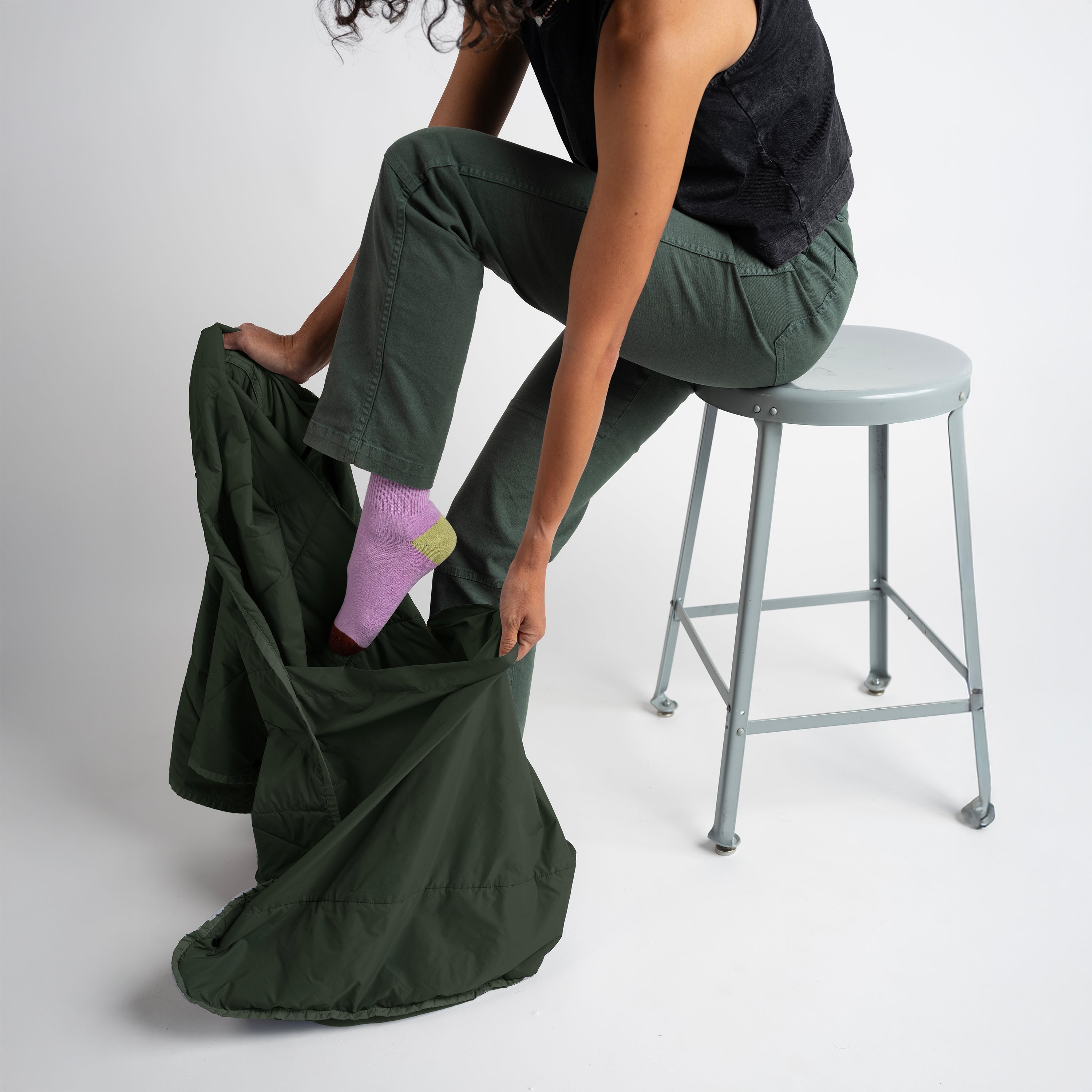 Person sitting on a stool with a large green bag filled with items on a white background