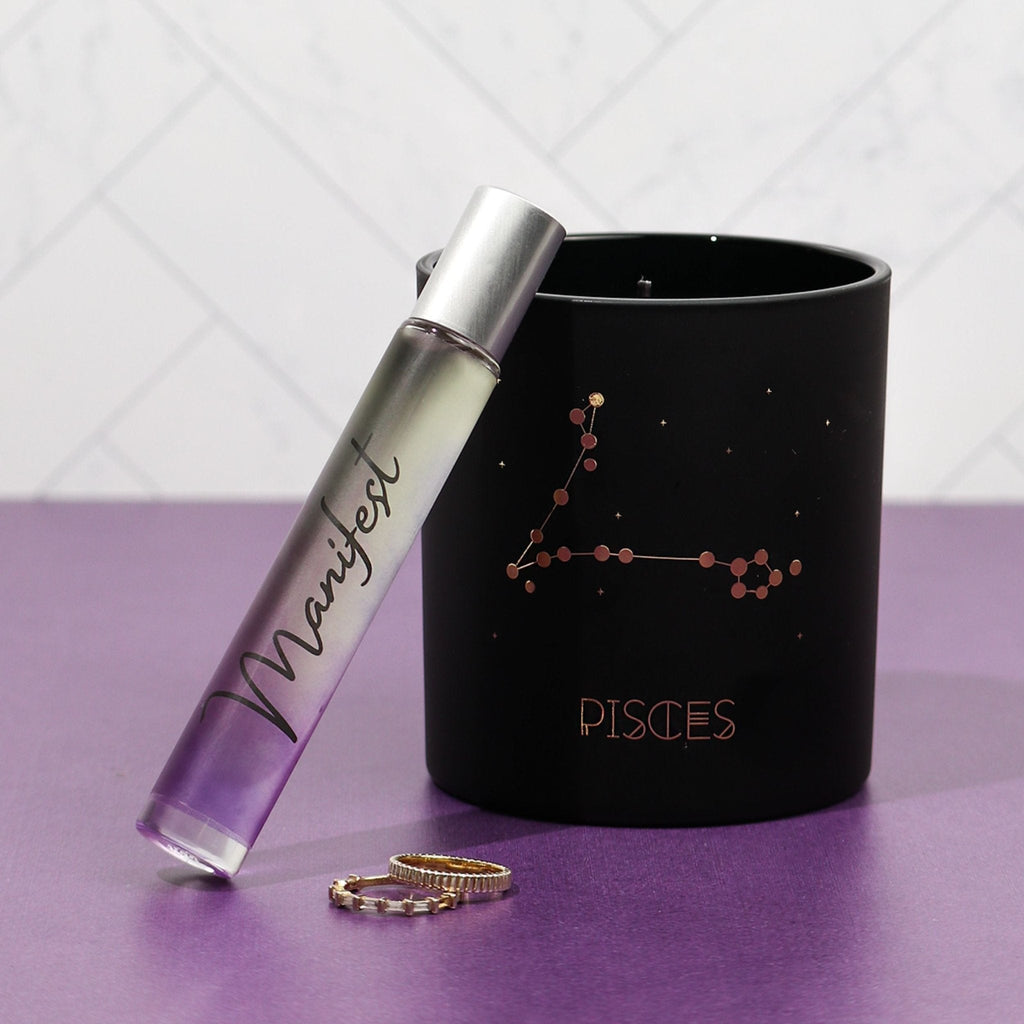 Black candle with 'Pisces' design and 'Manifest' rollerball on a purple surface.