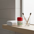 Red and white container of travel-sized Old Spice Fiji Deodorant on a bathroom shelf with a glass of toothbrushes and a towel.