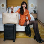 Woman in an orange dress holding a beige handbag with a black suitcase and small black bag on a bed.