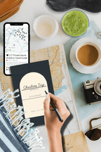 Hand writing in a black Everywhere travel planner beside a phone, coffee and camera